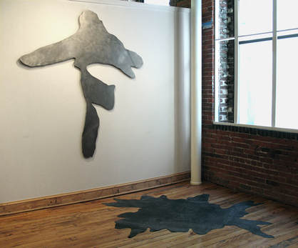 In the Beland Gallery in Lawrence, MA, a wall hung tree footprint made of steel and on the floor another tree footprint made of black roofing rubber. 