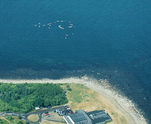 Aerial view of thirty sea kayakers making a spiral on a bay and a large stone spiral on land to reflect the way global ocean currents flow.