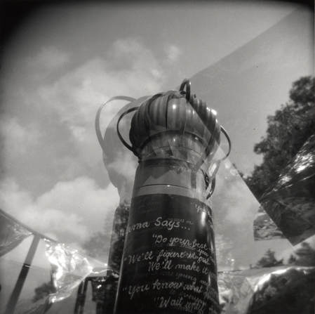 Square, black and white double exposure Holga photo of part of Mama Says showing the vent fan top against the sky, a tree and wavy mylar sheets moving in breeze.
