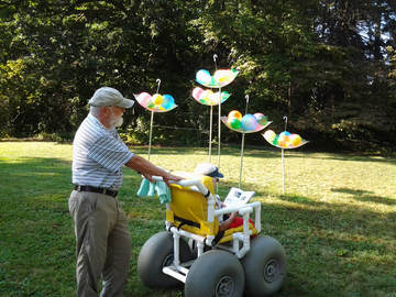 A man pushing an elderly woman in an all-terrain wheelchair with balloon tires at the Maudslay Outdoor Sculpture exhibit at a state park. An installation of white upside-down umbrellas filled with big, pastel colored balls is in the background.