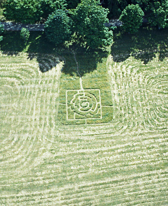 Aerial view of a maze cut into a field near a row of trees and a pathway. The mowing pattern of the field around the maze.