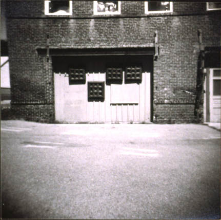 A black and white Holga photograph of the back of the former Amesbury Artworks building with art on the wall and eerie reflected light  from the windowson the pavement. 