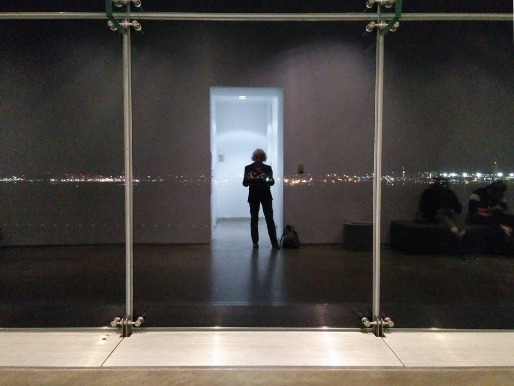 A self portrait at the ICA in Boston showing me silhouetted in a blue doorway, surrounded by darker walls. A horizontal stripe of city lights and two vertical supports of the glass windows are superimposed. It is a glass reflection.