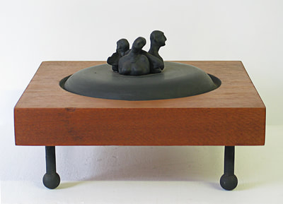 A brown, square wooden shape wiht four black legs and atop the center is a round black shape from which appear the tops of three figures - a woman and two men - and between them, a fish.