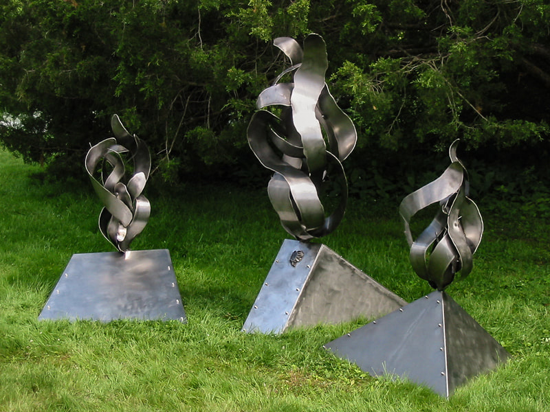 Against lawn and evergreens of the Moses-Kent House Museum three steel shapes rest. Their bases are geometric and atop each are organic flame or wind-like shapes curling and twisting upward.
