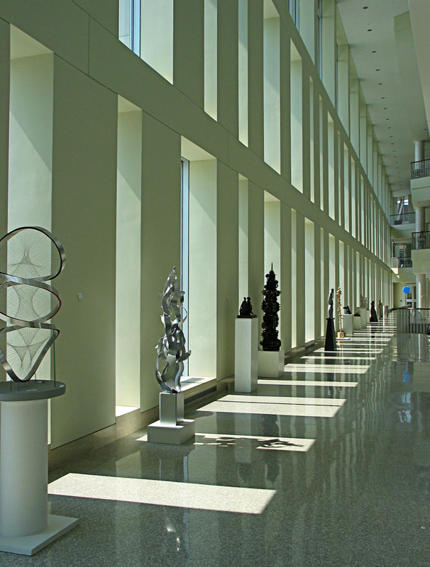 Kinetic Forces Upright shown with other scultpures against a row of windows in a long architectural environment.