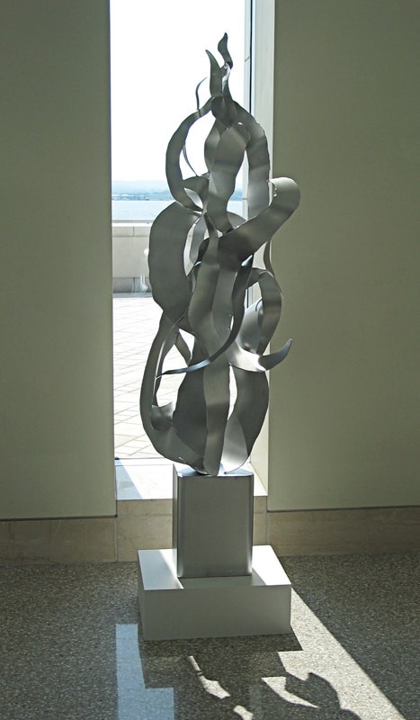 Silhouetted against a window at U Mass Boston, the silvery steel sculpture has a rectangular bottom from which organic shapes curl and twist upward like fire or smoke.