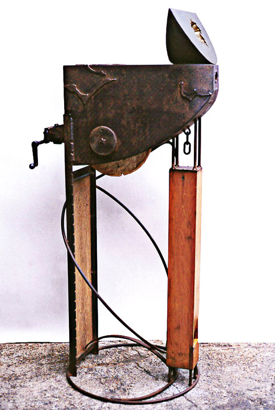A wedge shape is on a larger body supported by a steel panel in back and a wooden organ pipe in front over a round base. In back is a handle that can be turned and a wheel to rotate.