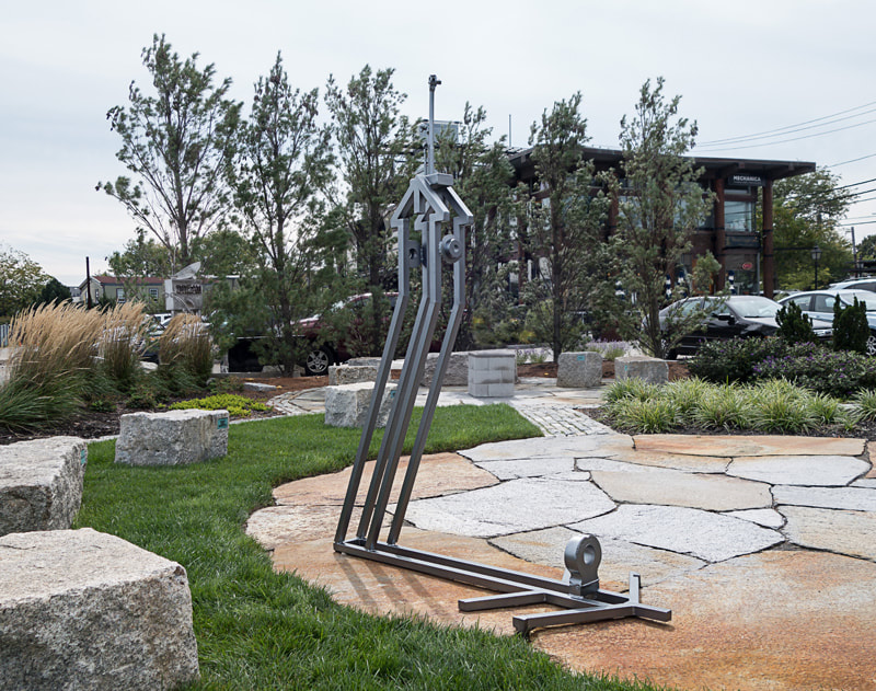 Outdoors in a sculpture garden partially paved with granite slabs, is tall silvery steel Perseverance. A home shape is topped by a family of wrenches and below is the shadow supporting the sculpture.