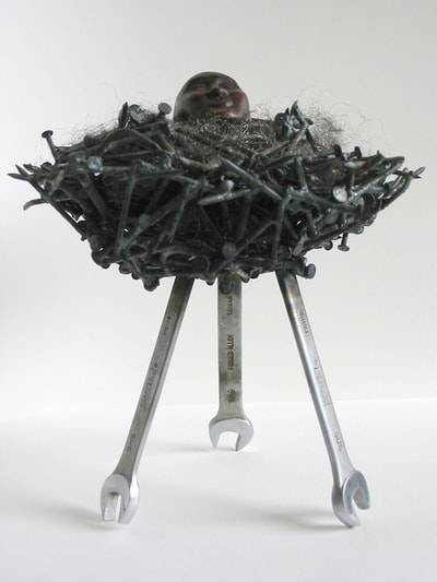 A nest of nails supported by three wrenches as legs. In the nest is a hydrocal egg with a female face on one side and a male on the other. 