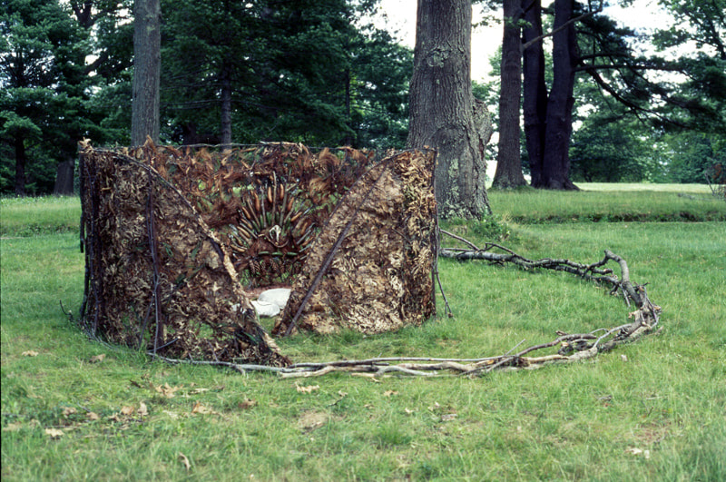 In the forest a spiral shaped enclosure with walls of leaves, sticks and pinecones arranged in a tribal pattern. Small white cushoins are inside. 