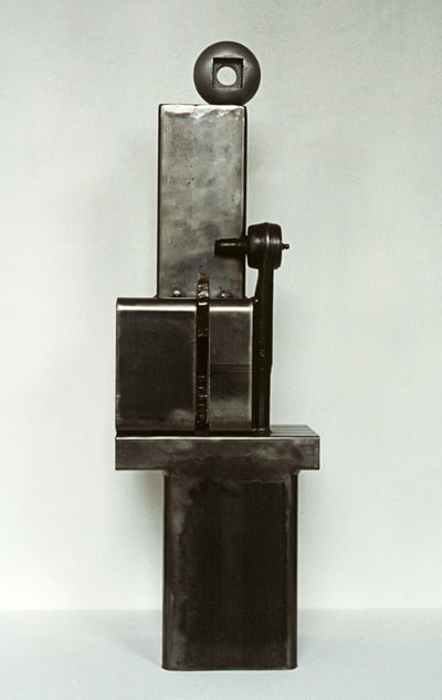 A dark steel vertical sculpture of square shapes with a discarded tool and on top a cast aluminum sphere-like shape.
