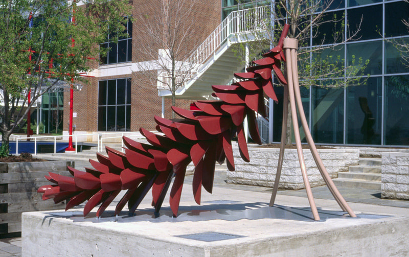 8.5' long red steel pinecone held up on one end by 5 ocher colored pipes on a grey shadow. City setting.