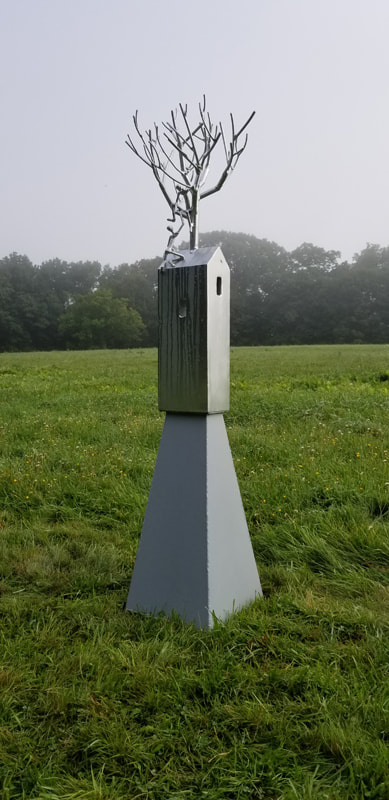 Another view by David Davies of Symbiotic steel sculpture  on a foggy day. There is a base with a tall, home-like shape and a tree on top. it is in a field with trees in the distance.